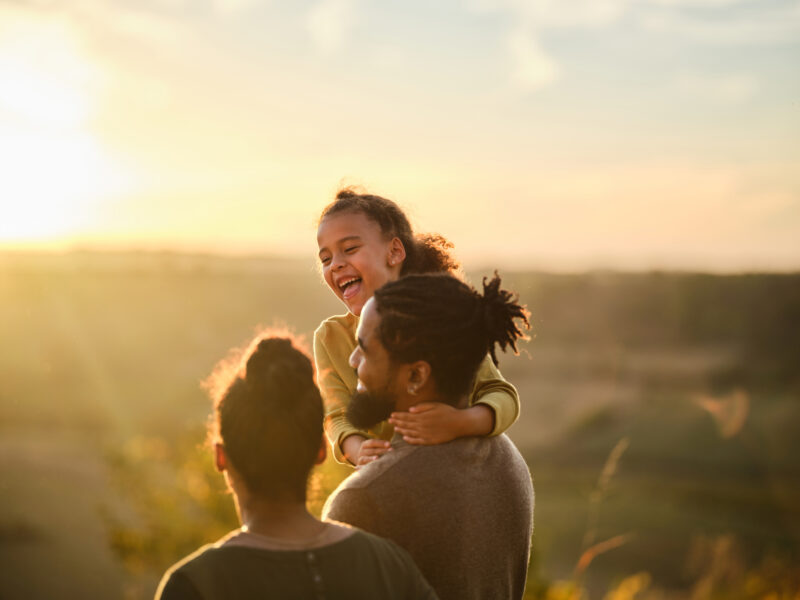A young girl smiles and laughs while being carried on a man's shoulders, with another adult standing nearby, symbolizing the support and care that can be essential when helping a loved one struggling with alcoholism.