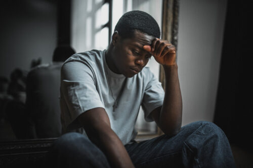 Young Black man sitting by a window with his head resting on his hand, looking overwhelmed or stressed. This image reflects the emotional weight of high cortisol levels and the connection to anxiety, as explained by therapists.