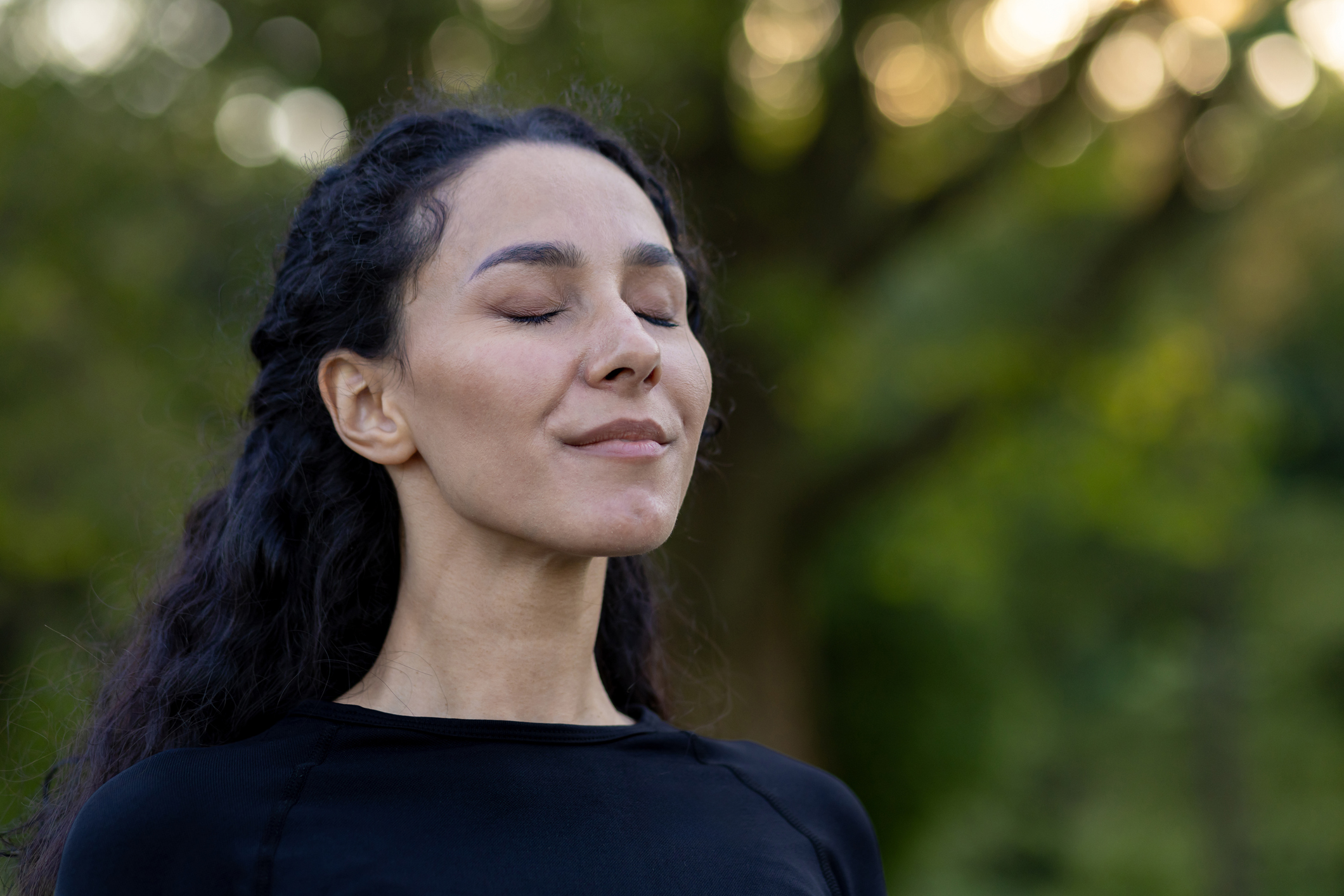 A woman with dark curly hair stands outdoors with her eyes closed and a calm smile on her face. She appears peaceful and grounded, symbolizing emotional balance and healthy coping strategies after quitting smoking.