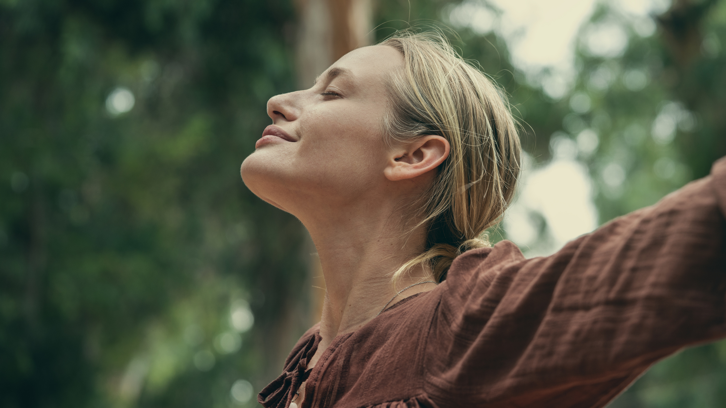 A close-up profile of a blonde woman standing outdoors with her eyes closed and a serene smile. Her head is tilted slightly upward, appearing relaxed and at peace, with a soft-focus background of trees.