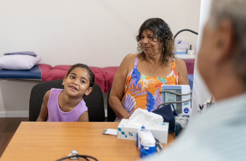 A young girl smiles while sitting beside an older woman during a medical appointment, with a blood pressure monitor and tissues on the table, illustrating the topic of finding the best medication for children with ADHD and ODD.