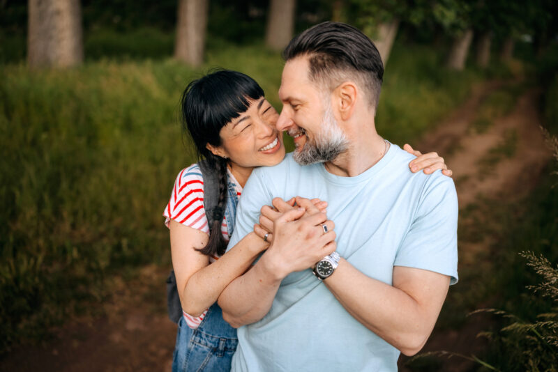 A joyful couple stands outdoors, smiling and holding hands. The woman, wearing a striped shirt and overalls, embraces the man from behind. They gaze at each other affectionately, surrounded by greenery and soft natural light.