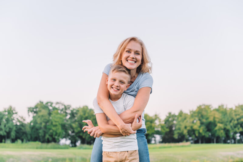 Smiling woman embraces a young boy from behind in a warm, supportive hug outdoors, representing the importance of connection and reassurance in helping kids manage anxiety.