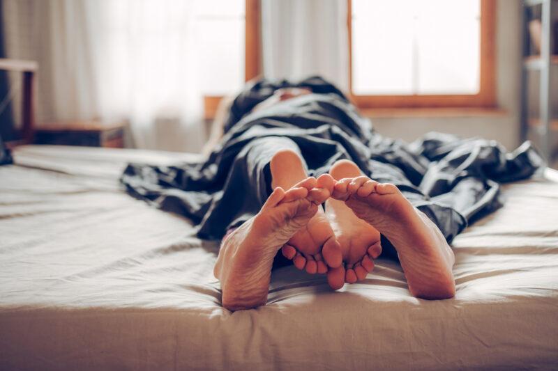 A close-up of two pairs of feet intertwined under a blanket, lying in bed. The focus is on the feet, with the rest of the bodies blurred in the background. Morning light filters through nearby windows, suggesting a quiet, intimate setting.