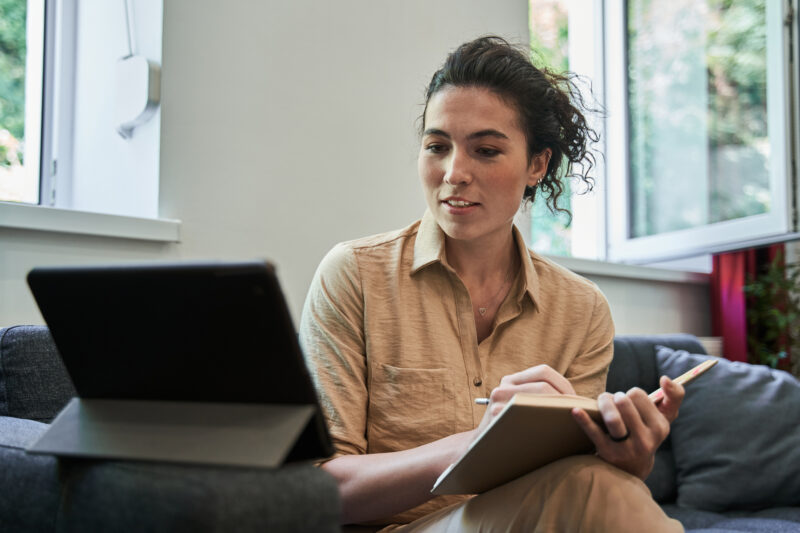 A young woman sitting with a notebook, engaged in an online therapy session on her tablet, representing CBT for eating disorders.