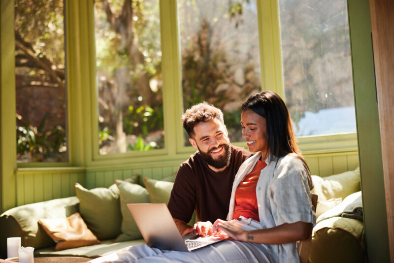 A smiling couple sitting close together on a couch with a laptop, illustrating a temporary calm and connection during the honeymoon stage of the cycle of abuse.