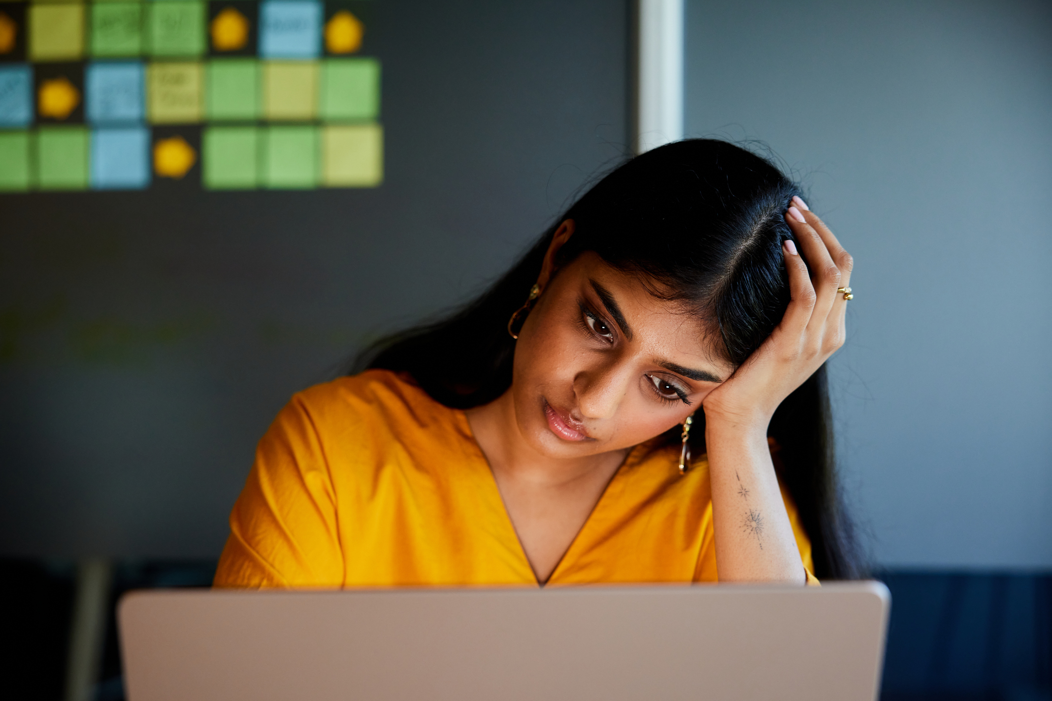 Stressed woman at work holding head while working on laptop, symbolizing workplace stress management challenges.