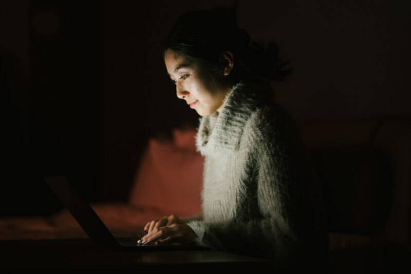 Young woman attending virtual evening IOP session from home, lying comfortably in bed with laptop open.