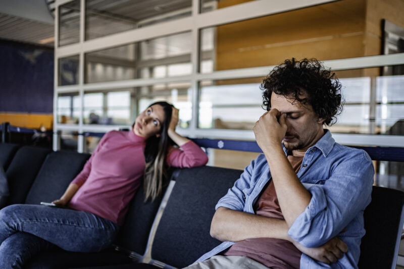 A distressed couple sitting apart in a waiting area, both showing signs of emotional exhaustion and frustration — representing the tension and conflict stage in the cycle of abuse.