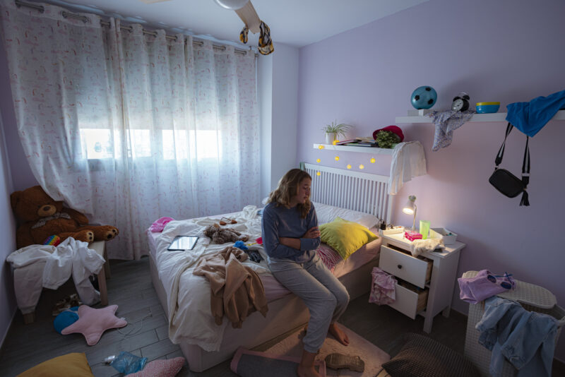 A person sitting on the edge of their unmade bed in a dimly lit, cluttered bedroom, hugging themselves and looking away, conveying feelings of isolation and emotional distance.