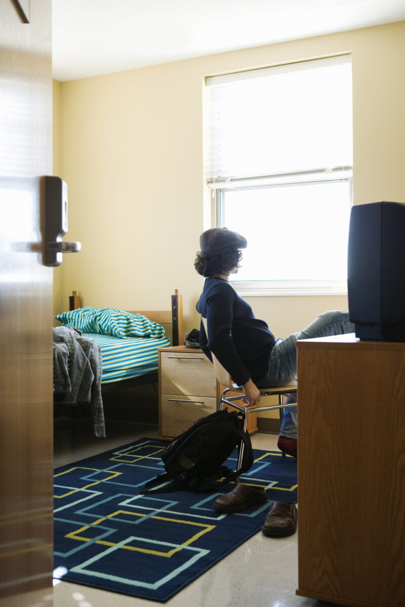 A college student sitting alone in a dorm room, leaning back in a chair and staring out the window with a distant expression, conveying feelings of isolation and depression.