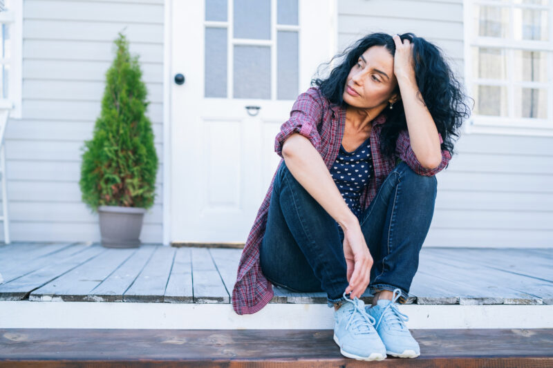 A woman sits on her porch with her head resting on her hand, appearing reflective and somber, symbolizing the emotional process of healing from trauma.