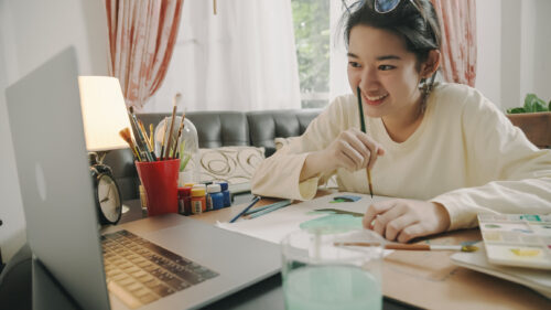 Teenage girl hand drawing and painting her art in the living room during a Charlie Health virtual session. She is using a laptop having a Video call with members of her peer group while using watercolor and brush to create art project in to white paper form her imagination.