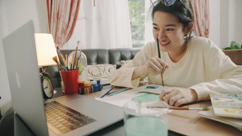 Teenage girl hand drawing and painting her art in the living room during a Charlie Health virtual session. She is using a laptop having a Video call with members of her peer group while using watercolor and brush to create art project in to white paper form her imagination.