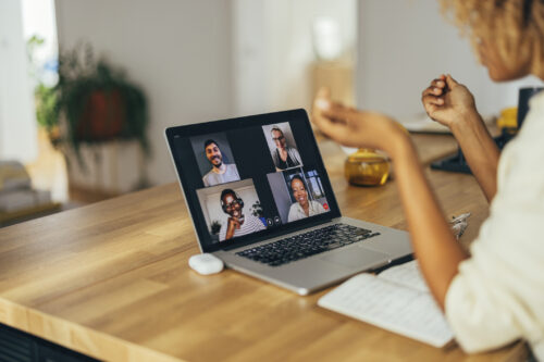 American woman is sitting at a desk and having a video call on a laptop with a group of four people, two males, and two females who are part of her Charlie Health alumni group. She is explaining something to them and having her notes in front of her. She has glasses, a glass of water, and a headphones case on the desk. She might be in an office or working from home.
