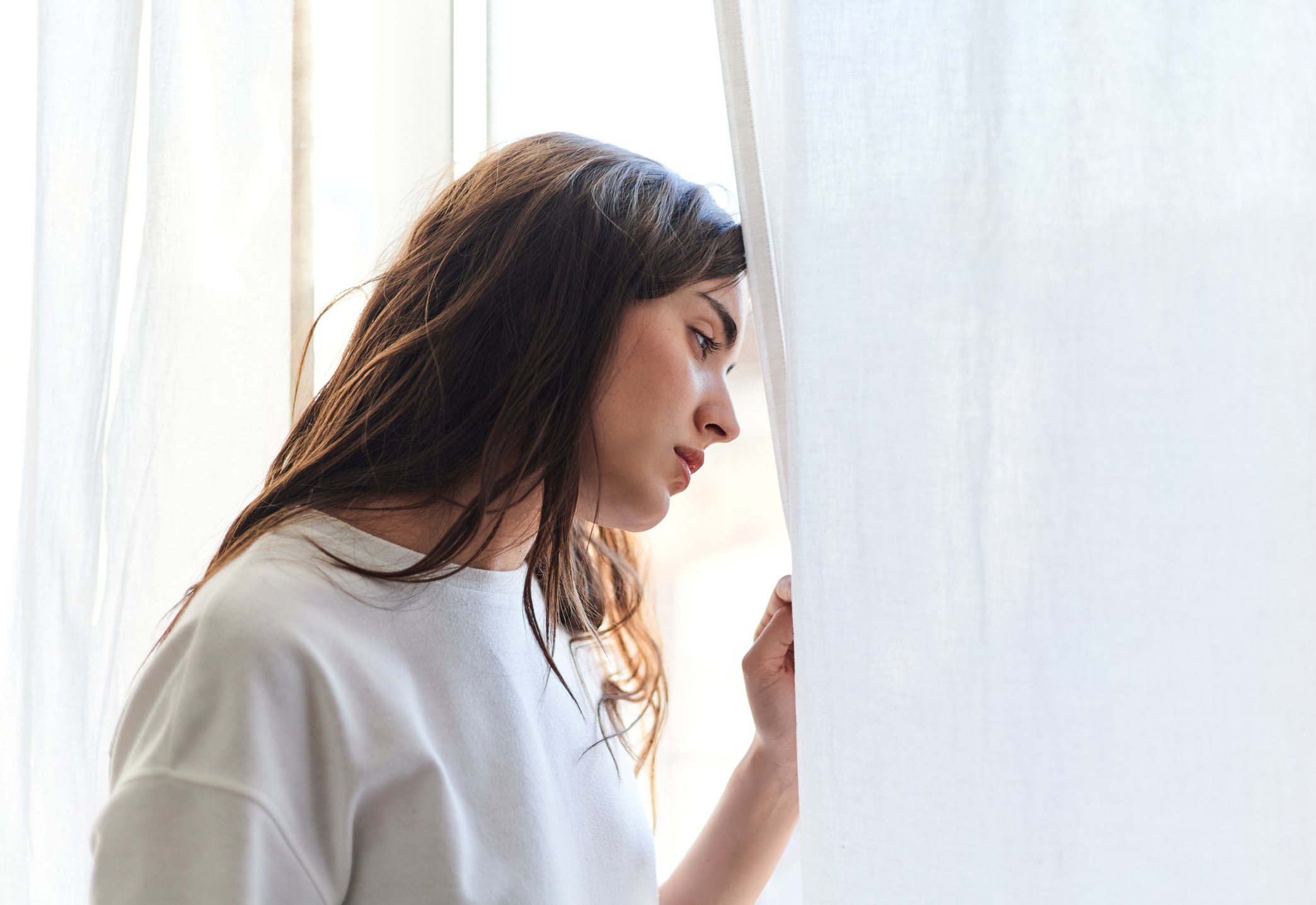 A young woman stands by a window looking out pensively, reflecting feelings of loneliness and longing often associated with separation anxiety.