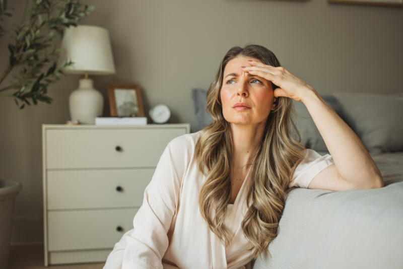 A middle-aged woman sits on a couch with her hand on her forehead, appearing thoughtful and concerned, reflecting on the challenges of managing ADHD symptoms during menopause.