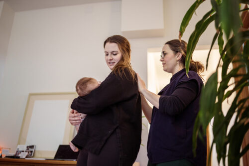 A postpartum doula gently supports a new mother holding her baby in a calm home setting, offering guidance and reassurance during the early weeks of parenthood.