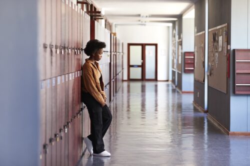 Side view of teenage girl leaning on lockers while standing in hallway at high school, contemplating her anxiety and mental health.