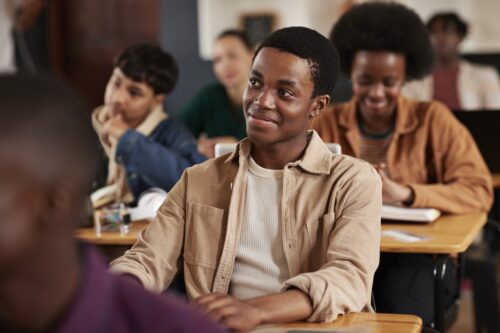 Smiling teenage boy sitting at desk in high school classroom, among his friends, developing a healthy relationship with social media.