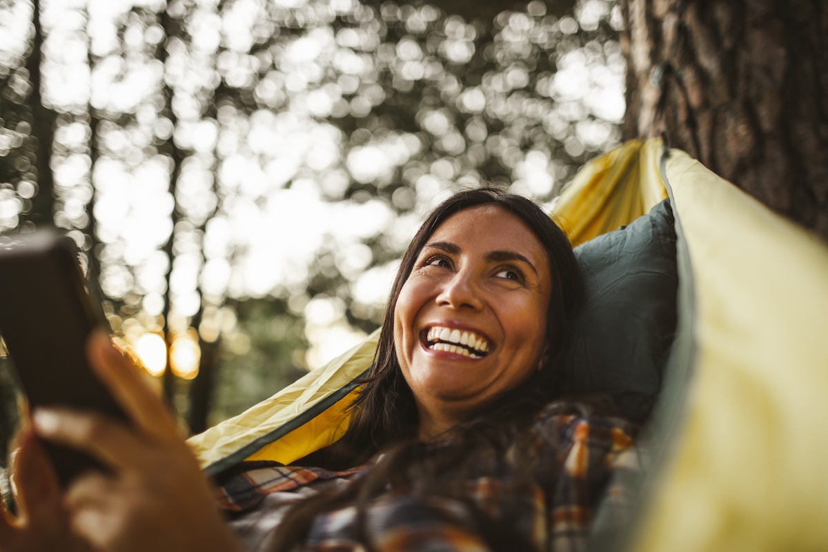 Smiling woman using smart phone while lying over hammock in forest.